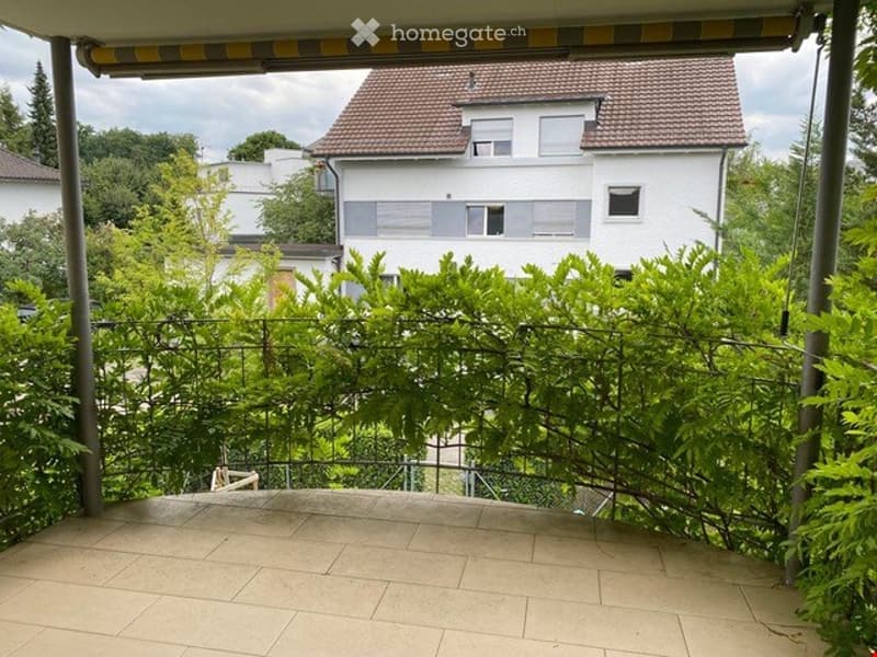 Balcony with tiled floor, metal railing, and lush greenery, overlooking a residential area with houses and trees.
