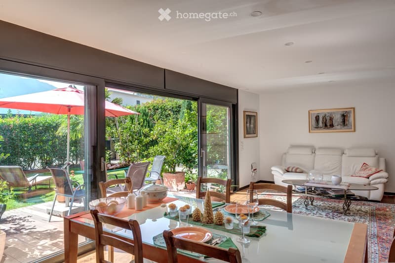 Bright living and dining area with large glass doors opening to a garden, featuring a white sofa, coffee table, dining table with chairs, and decorative items.