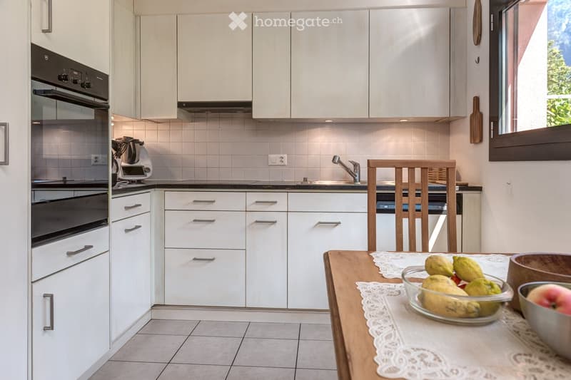 Modern kitchen with white cabinets, stainless steel appliances, a wooden dining table with a lace tablecloth, and a bowl of fruit on the table.