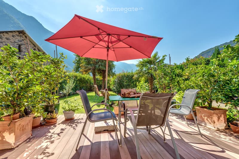 A wooden deck with a table, chairs, and an umbrella, surrounded by potted plants and a scenic mountain view.