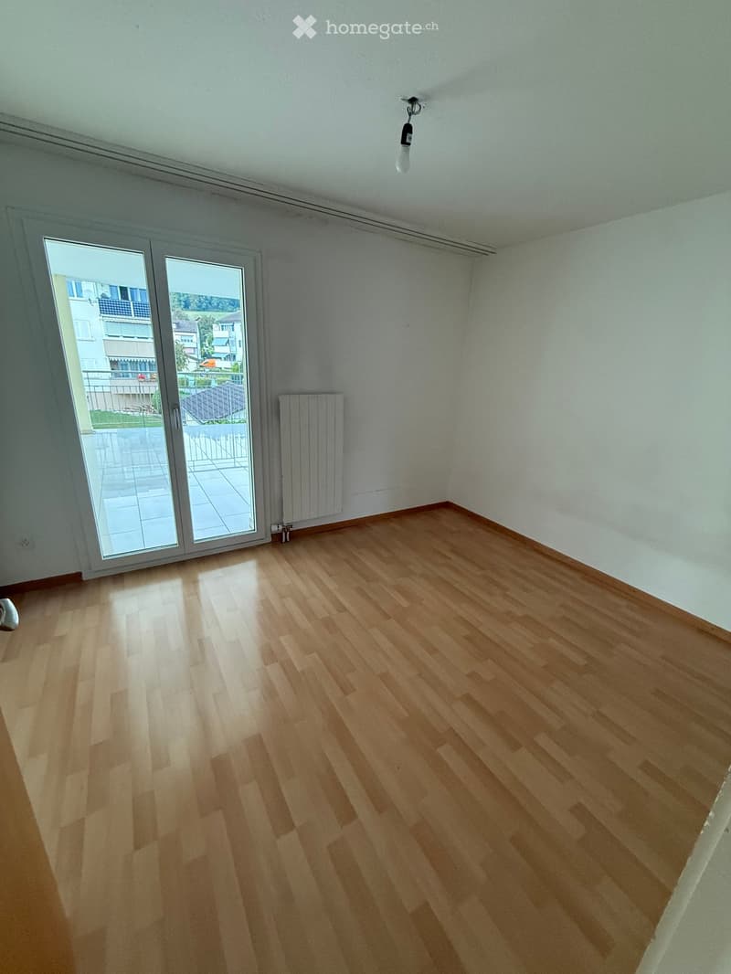 Empty room with wooden flooring, white walls, large sliding glass door leading to a balcony, radiator, and a single light fixture on the ceiling.