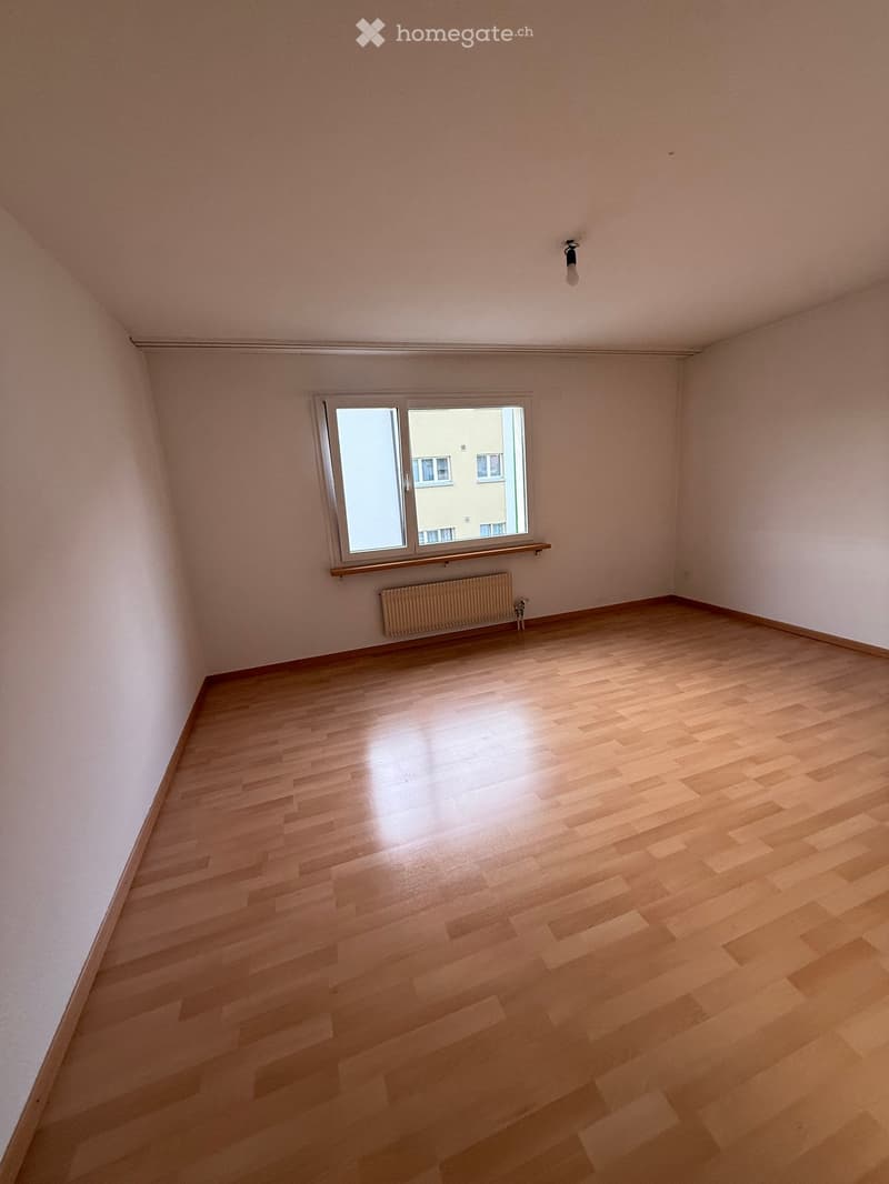 Empty room with light wooden flooring, white walls, and a single window with a radiator below it.