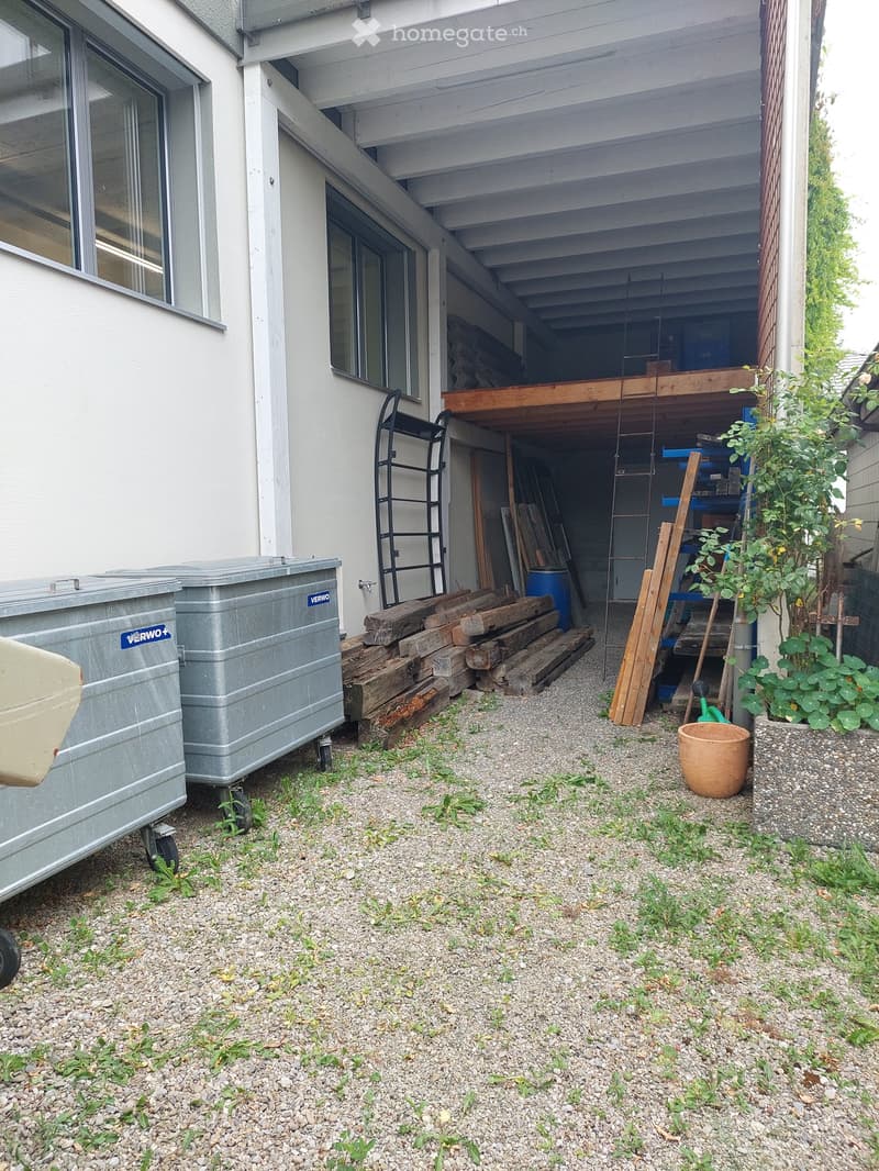 A storage area attached to a building with a gravel floor, two silver storage bins with wheels, a stack of wooden planks, a black ladder, a blue barrel, a blue shelving unit, a potted plant in a stone planter, and a brown bucket.