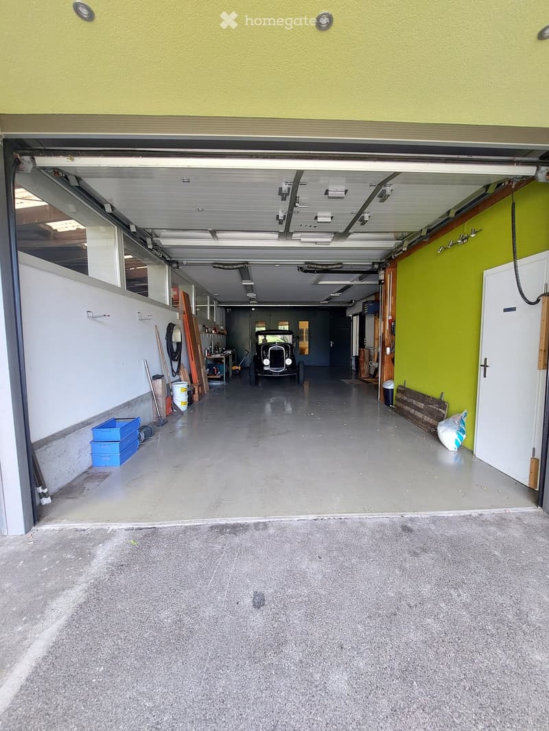 Garage with concrete floor, yellow walls, car parked, shelves, various tools and equipment, including a ladder, buckets, and a trolley, illuminated by white fluorescent lights