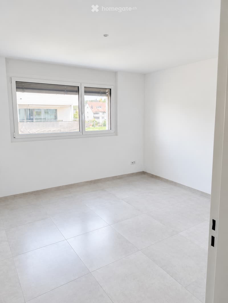 Empty room with white walls, tiled floor, and large windows offering a view of the outside area.