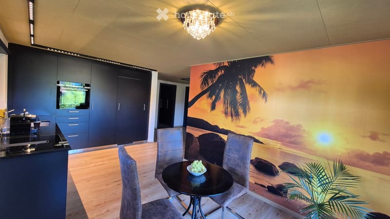 Modern kitchen with a dining area, featuring a black countertop, built-in cabinets, a stove, a refrigerator, a chandelier, and a round dining table with chairs. A large mural of a tropical beach decorates one wall.