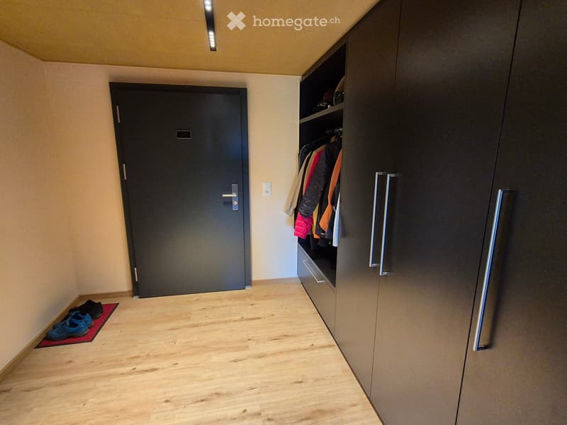 Clean hallway with wooden flooring, built-in closet with hanging clothes, a closed door, and a small red rug with shoes placed on it.