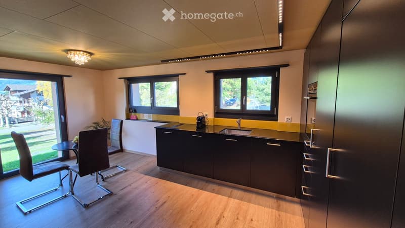 Modern kitchen with dark cabinets, a central island, and a small dining area with two chairs and a table. The space features large windows and a sleek design.