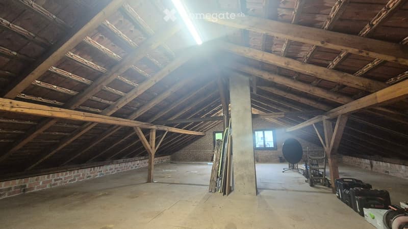 Empty attic space with exposed wooden beams and brick walls, concrete floor, and a single window letting in natural light.