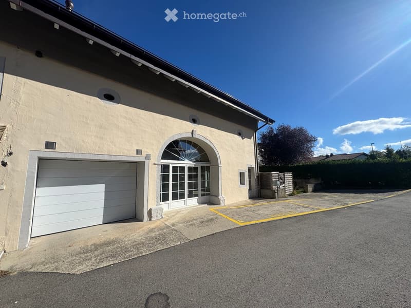 Single-story building with a garage and arched entrance, surrounded by a paved area with marked parking spaces.