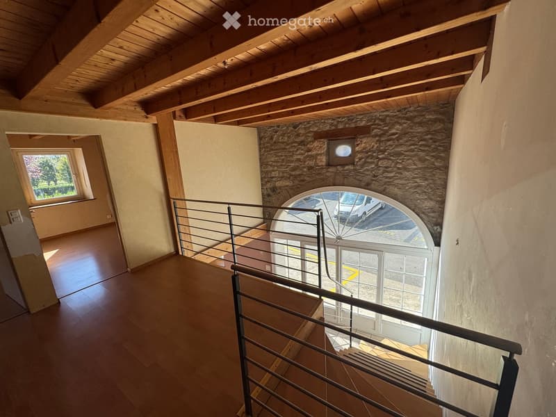 Spacious hallway with wooden flooring, exposed wooden beams on the ceiling, and a modern metal railing leading to a lower level. Features a large arched window and a smaller window on the left side.