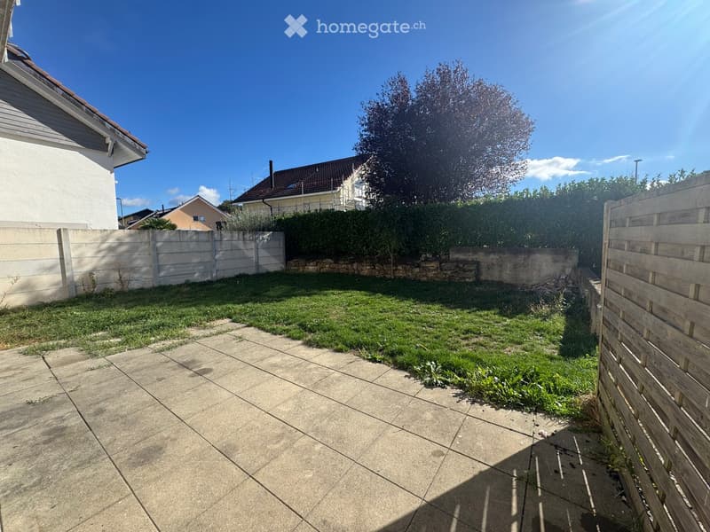 A small, well-maintained garden area with a concrete patio, grassy lawn, and surrounding wooden and concrete fences. The area is enclosed and features a clear blue sky and neighboring houses in the background.