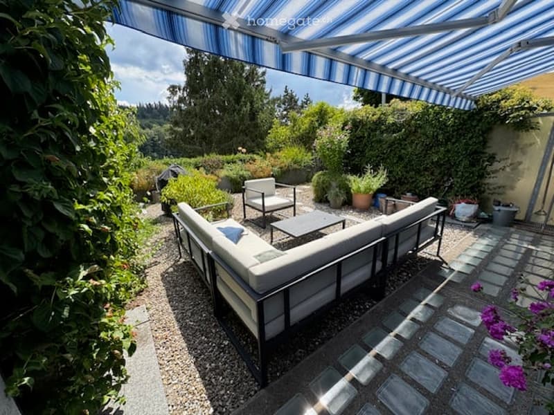 A covered outdoor patio with a white sectional sofa, a coffee table, and a couple of armchairs, surrounded by lush greenery and potted plants.