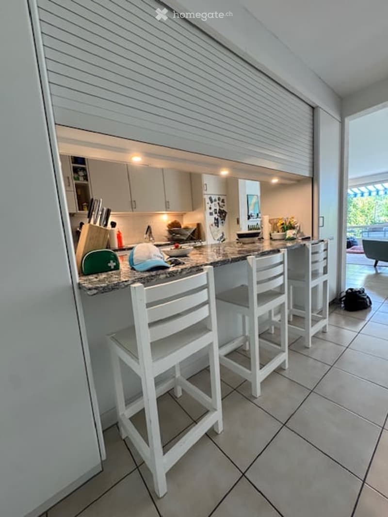 Modern kitchen with a bar counter, white stools, and a view to a balcony. The counter features a knife block, a bowl, a vase with flowers, and a mirror. The kitchen cabinets are white and sleek.