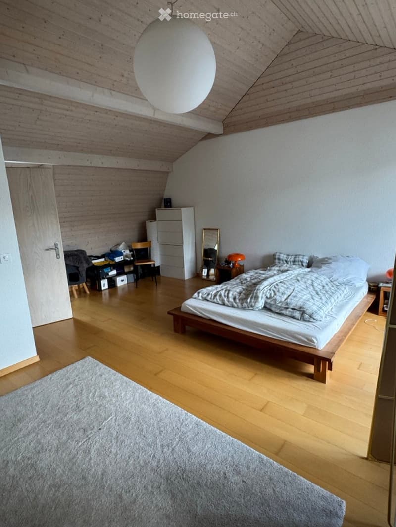 Cozy attic bedroom with wooden floors, slanted wooden ceiling, and a large round pendant light. Features a bed with a patterned duvet, a wooden chair, a small table with a lamp, a mirror, and a cabinet.