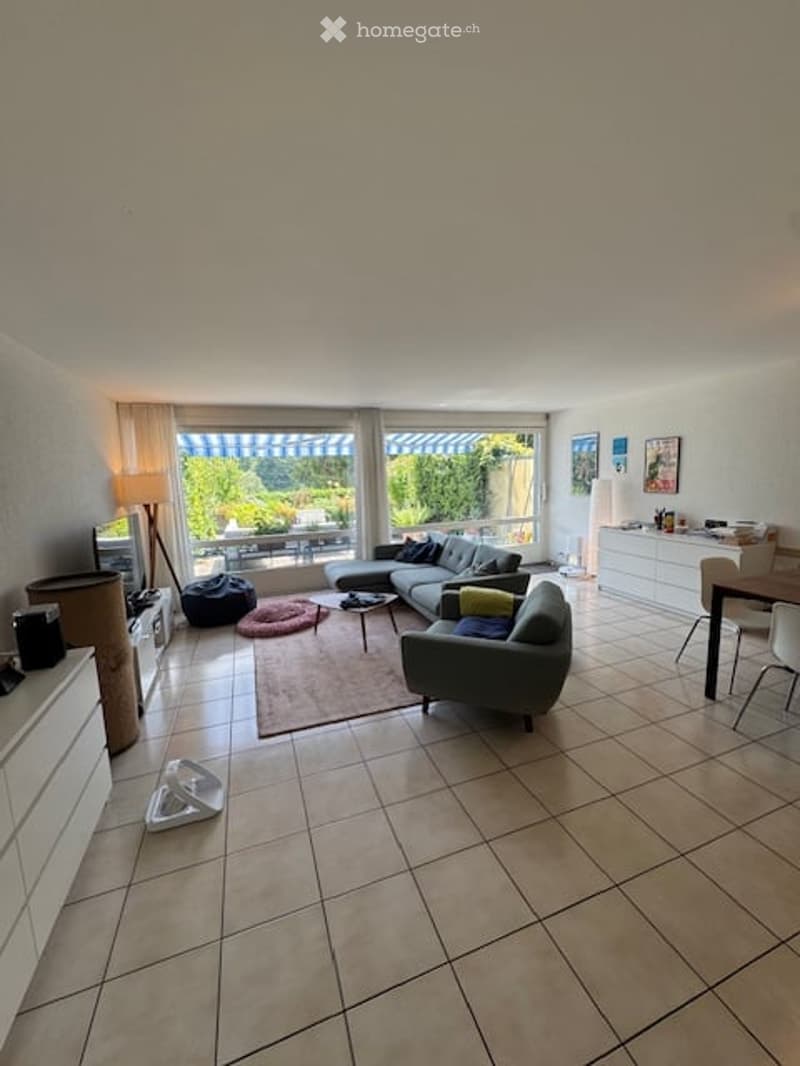 Bright living room with large windows offering a view, featuring a grey sectional sofa, coffee table, armchairs, floor lamp, TV stand, dining area with chairs and table, and white cabinets.