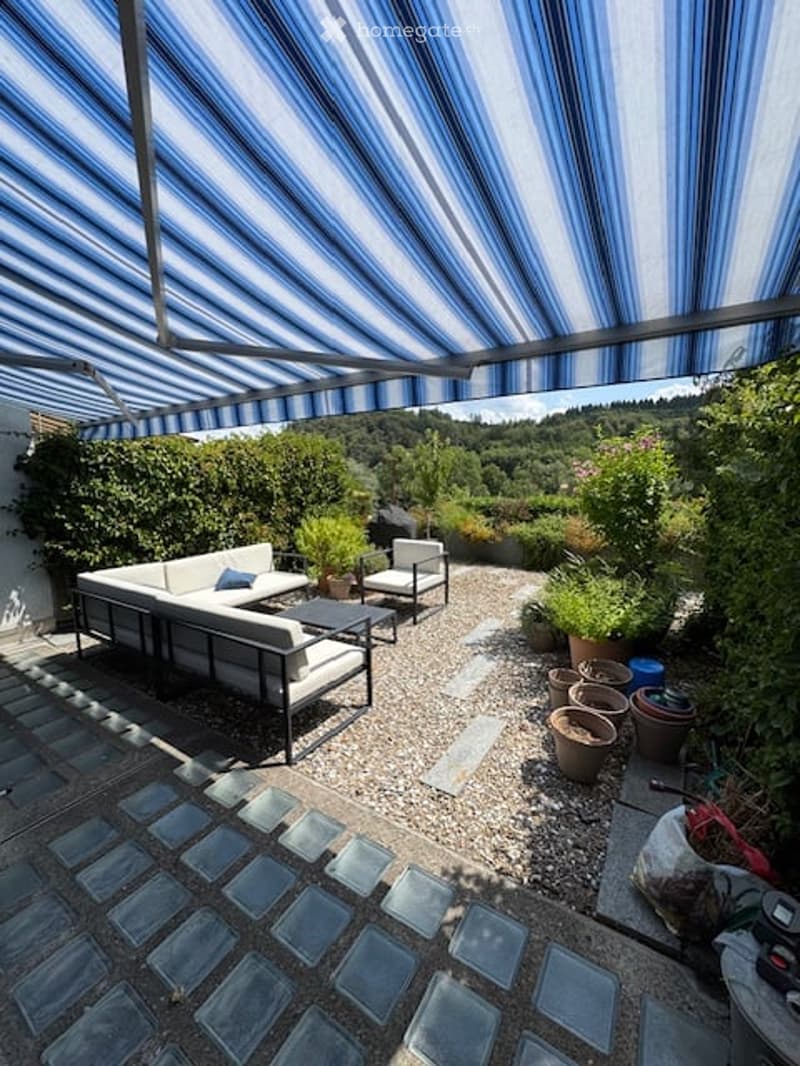 Outdoor patio area with a striped canopy, white sectional sofa, small coffee table, and potted plants. Gravel flooring and a view of greenery in the background.