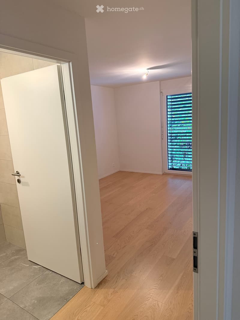 Empty room with light wooden flooring, white walls, and a large window with blinds. The room is connected to a hallway with a white door.