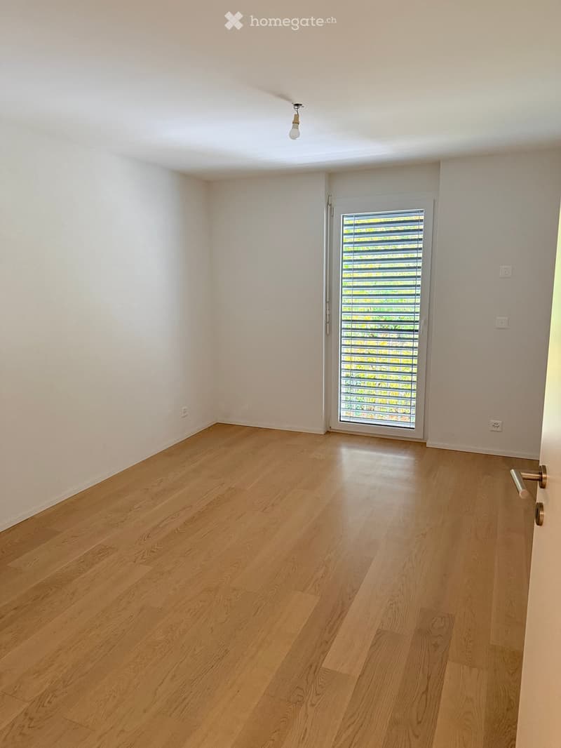 Empty room with light wooden flooring, white walls, and a sliding glass door with blinds.
