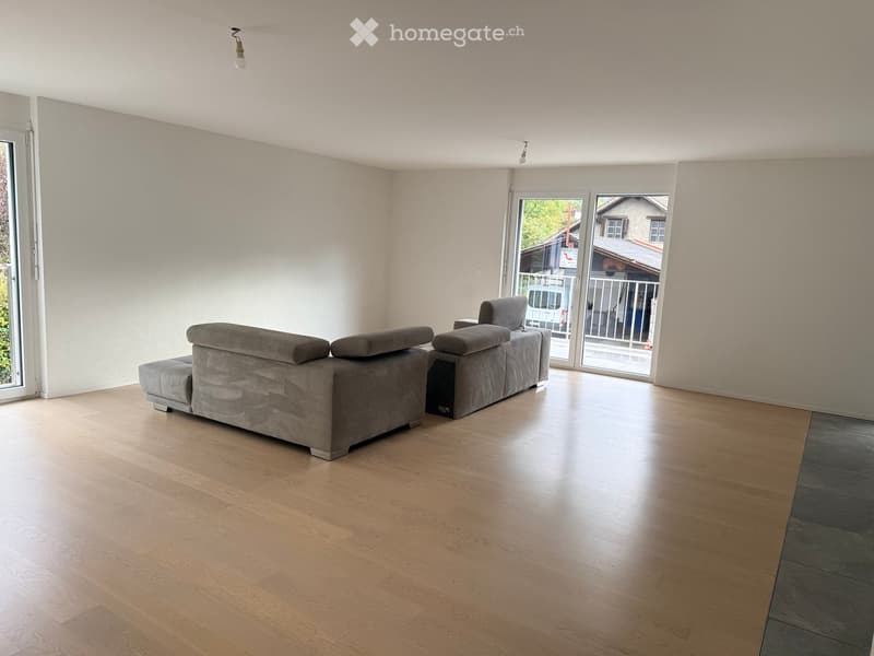 Modern living room with light wooden flooring, grey sectional sofa, large windows, and sliding glass doors leading to a balcony.