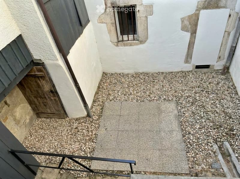 Small raised outdoor area with gravel and concrete steps, adjacent to a building with white walls and a wooden door.