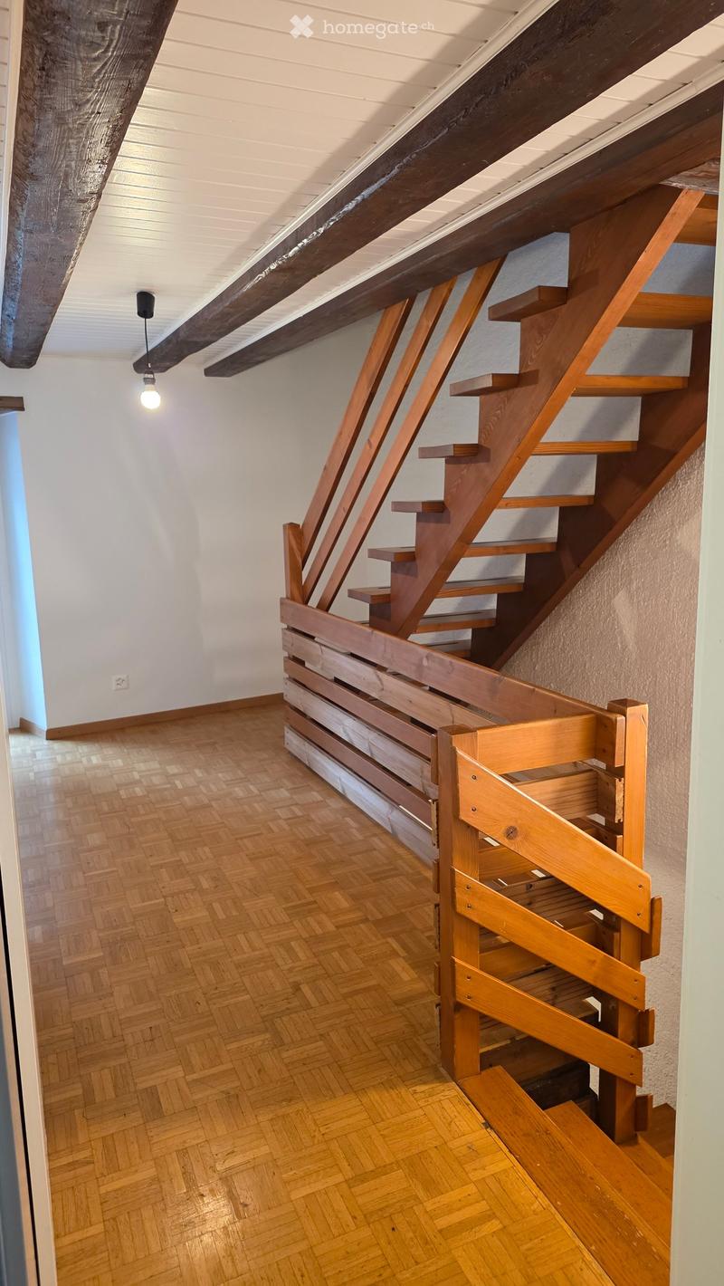 Wooden staircase with open wooden railings, wooden herringbone floor, white walls, exposed wooden beams on the ceiling, and a single hanging light fixture.