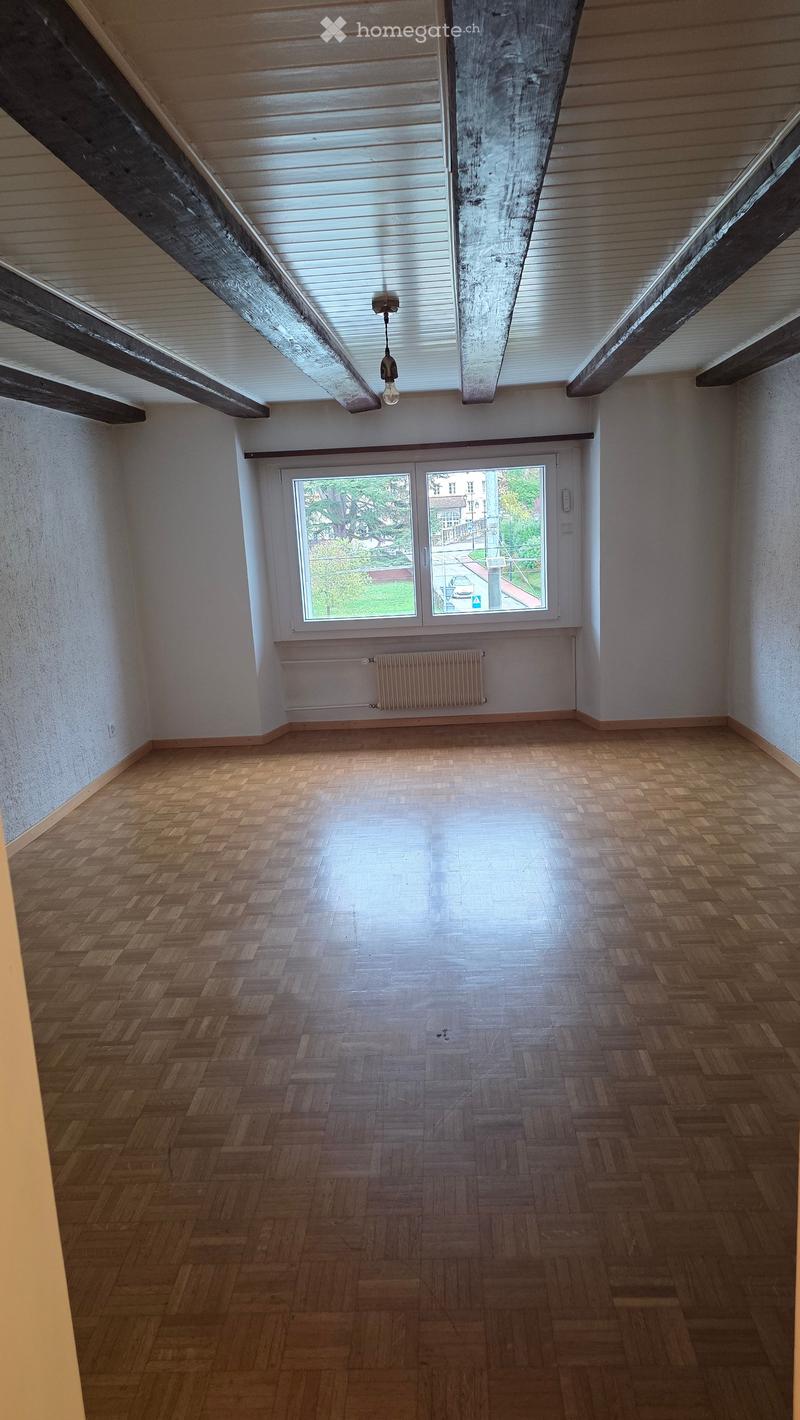 Empty room with wooden herringbone flooring, white walls, exposed wooden beams on the ceiling, and a large window with a view of the street.