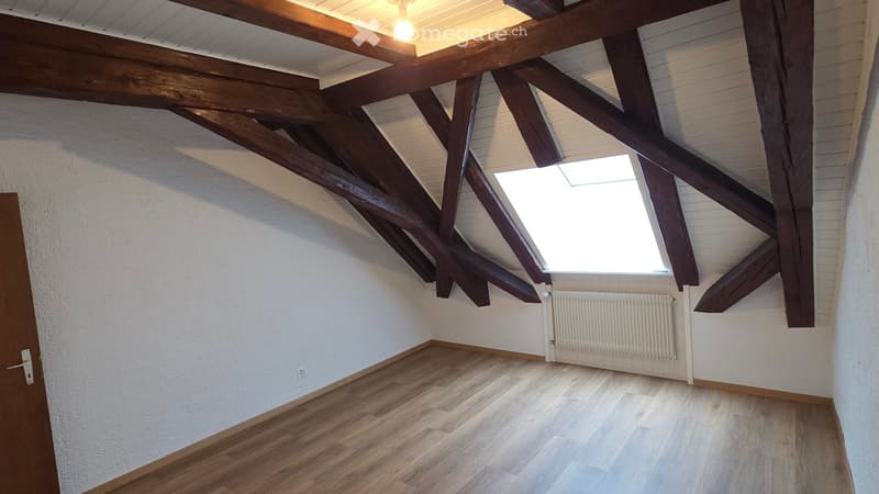Empty attic room with wooden beams, skylight window, wooden flooring, and white walls.