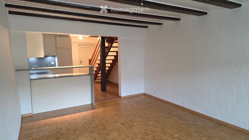 Empty room with wooden flooring, white walls, and a staircase leading to another area. There is a kitchen visible in the background with white cabinets and a countertop.