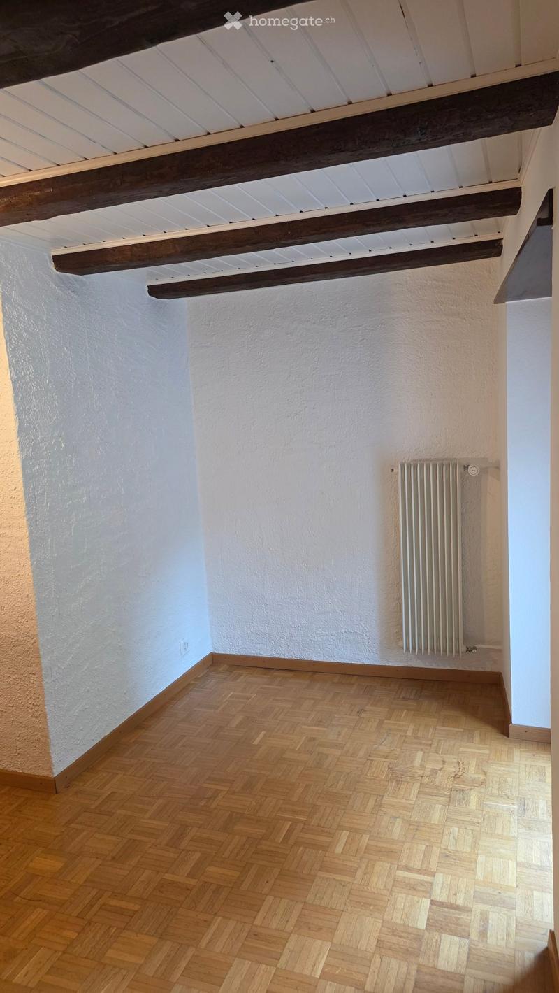 Empty room with wooden herringbone flooring, white walls, exposed wooden beams on the ceiling, and a radiator on one wall.