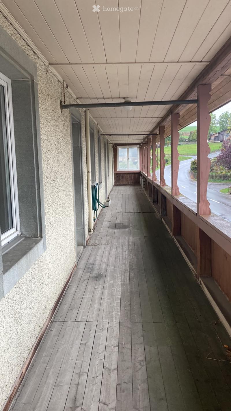 Wooden hallway with a covered walkway, featuring wooden flooring, white ceiling, and a view of the street and greenery outside.
