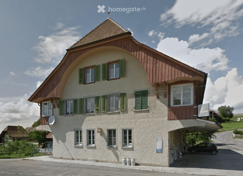 Traditional Swiss-style building with a pitched roof, green shutters, and a covered garage entrance.