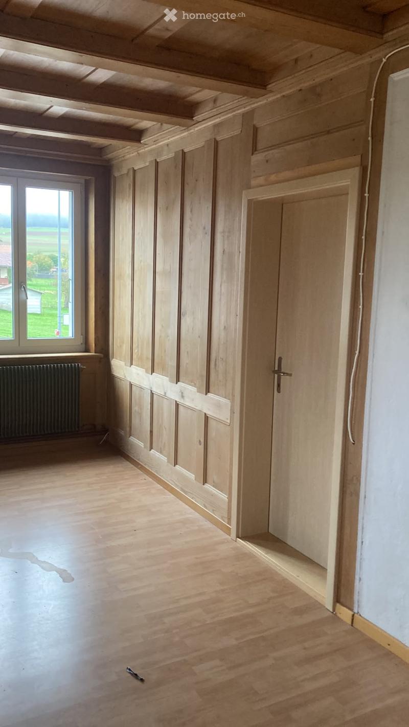 Empty room with wooden panel walls, exposed wooden ceiling beams, wooden floor, and a window with a view of the countryside.