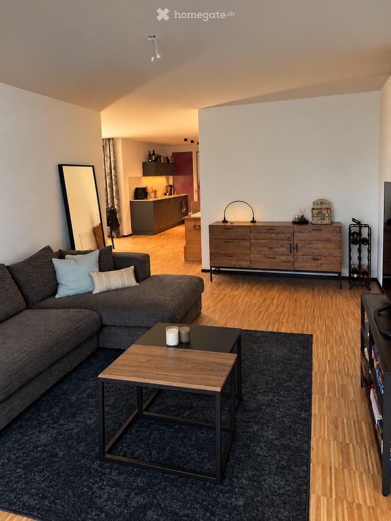 Modern living room with a sectional sofa, coffee table, large mirror, wooden dresser, wine rack, and a visible kitchen area with cabinets and appliances.