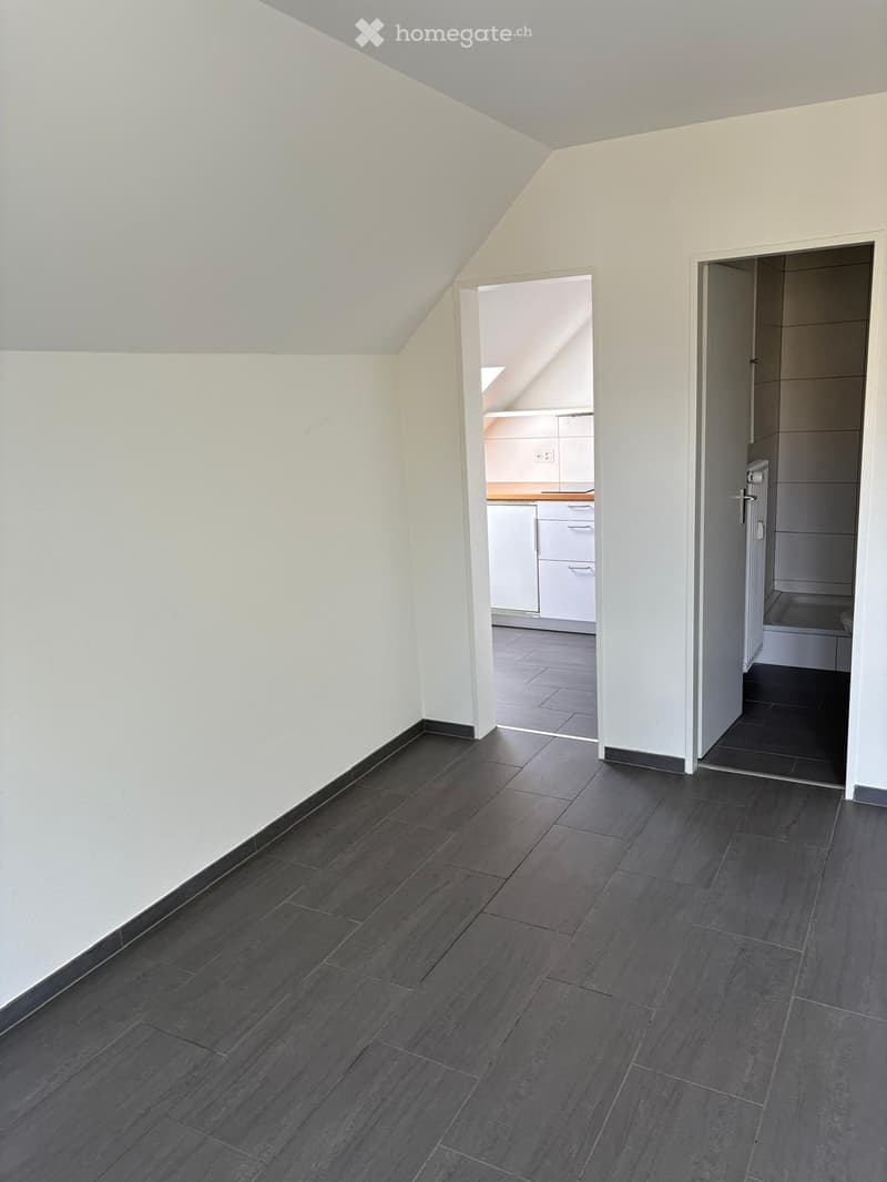 Empty room with dark grey tiled flooring and white walls, featuring an open doorway to a kitchen and another doorway to a bathroom.