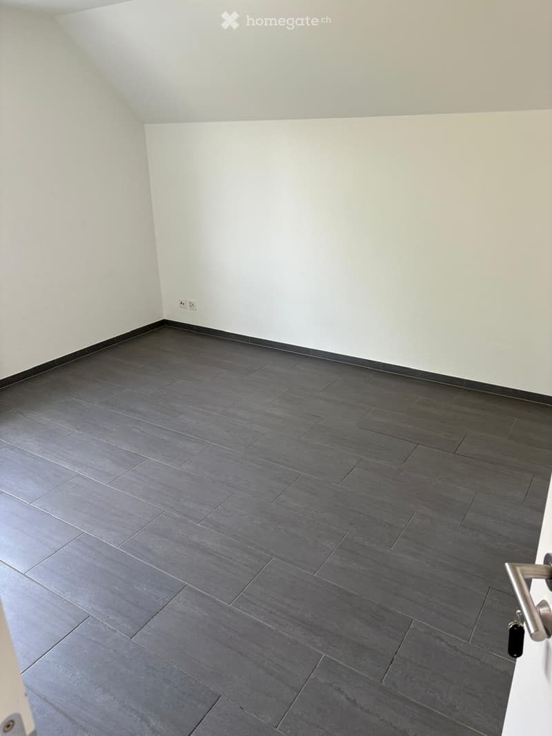 Empty room with grey tiled floor and white walls, featuring a door handle and electrical outlet visible.
