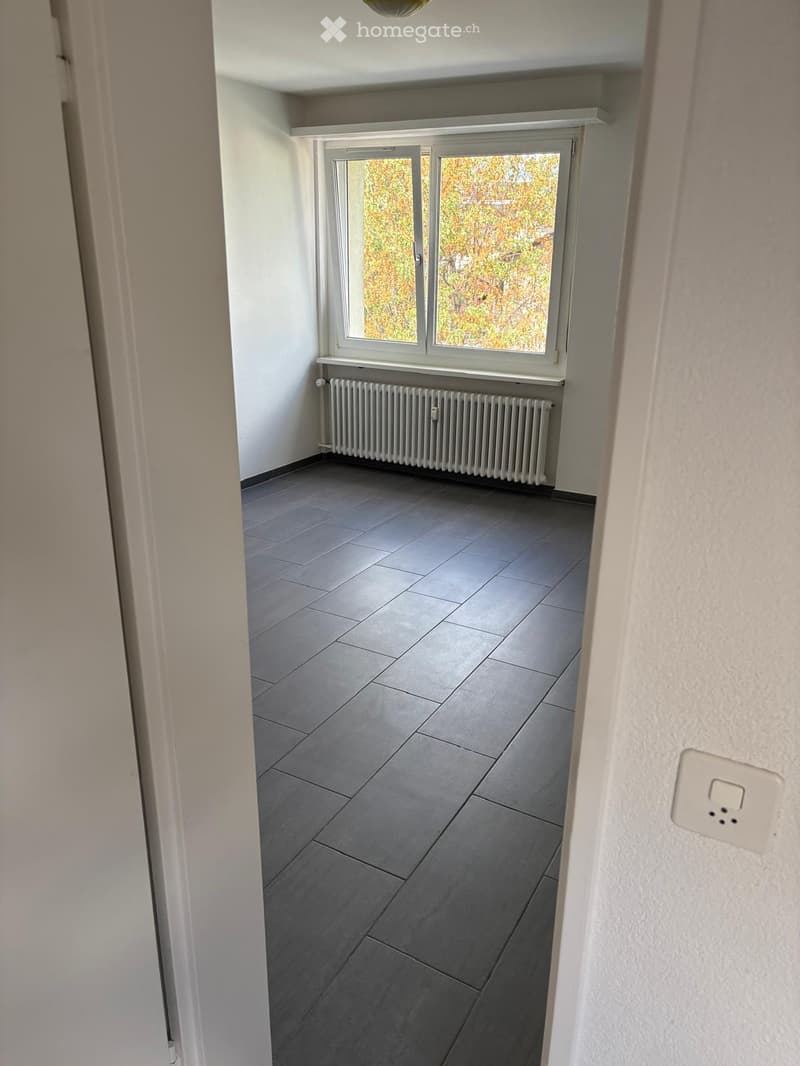 Empty room with grey tiled floor, white walls, large window with view of autumn foliage, radiator below window, and light switch on wall.