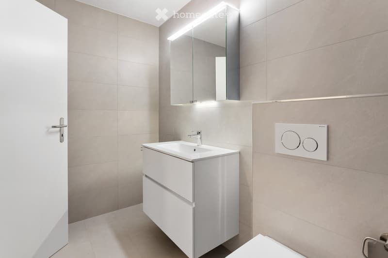 Modern bathroom with a white sink, faucet, mirrored cabinet with LED lighting, and a toilet. The walls and floor are tiled in a neutral beige color.
