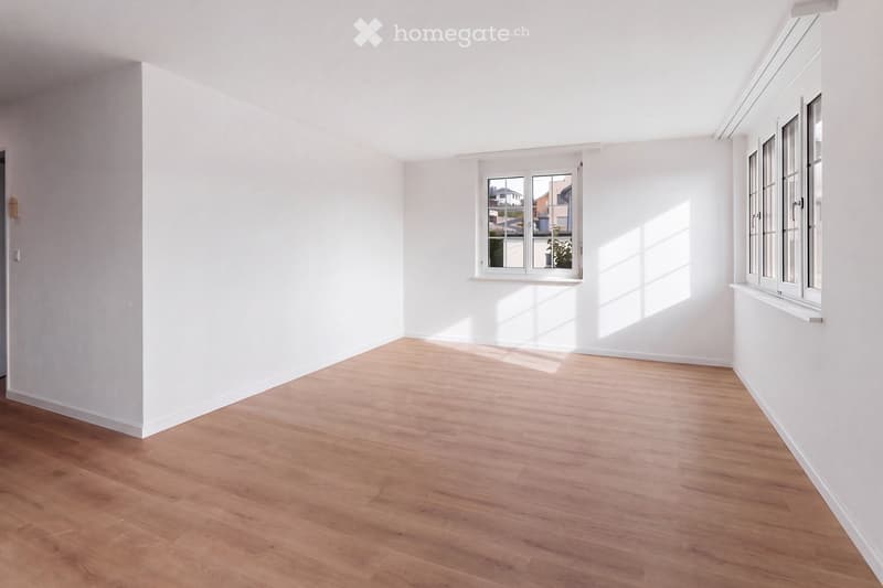Empty room with light wooden flooring and white walls, featuring two windows with white frames and a small plant visible outside one window.