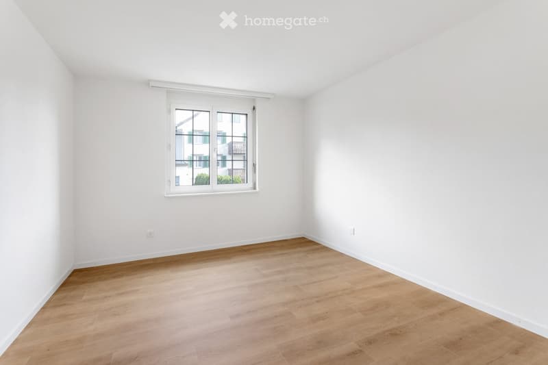 Empty room with white walls, wooden flooring, and a large window with white frame showing a view of a building and greenery outside.