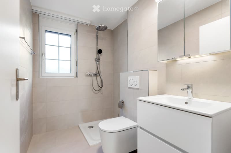 Modern bathroom with beige tiles, white fixtures, a toilet, sink, shower head, and a large mirror with built-in lighting.