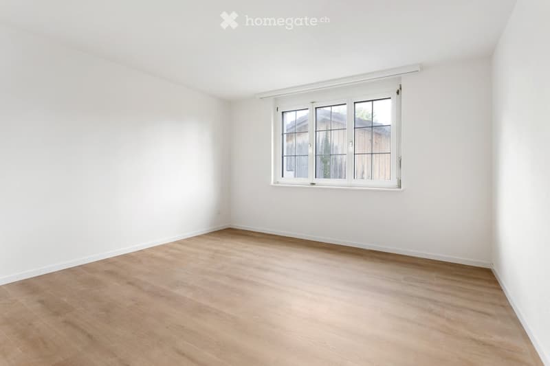 Empty room with light wooden flooring and white walls, featuring a large window with white frame and multiple panes.