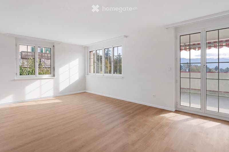 Empty room with light wooden flooring and white walls, featuring large windows and a sliding glass door leading to a balcony.