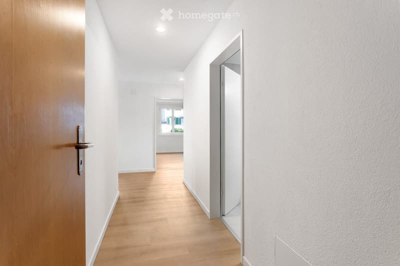 Clean, modern hallway with light wooden flooring, white walls, and recessed ceiling lighting. Features a wooden door on the left and an open doorway leading to another room on the right.