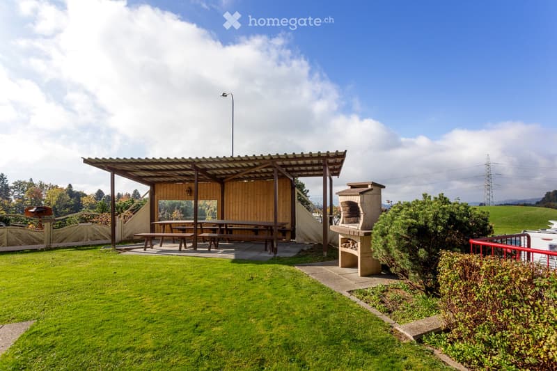 A well-maintained garden area with a wooden shelter, picnic tables, benches, and a stone pizza oven, surrounded by green grass and trees.