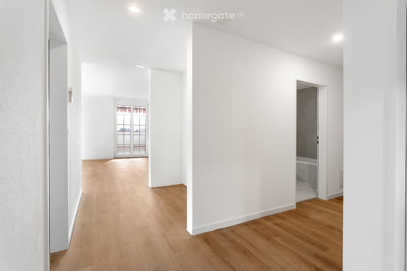 Bright, minimalist hallway with light wooden flooring, white walls, and recessed lighting. Features an open doorway leading to a room with a bathtub visible, and a glass door at the far end.