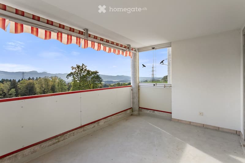 Empty balcony with white walls, red trim, and a retractable awning, offering a scenic view of trees and mountains.