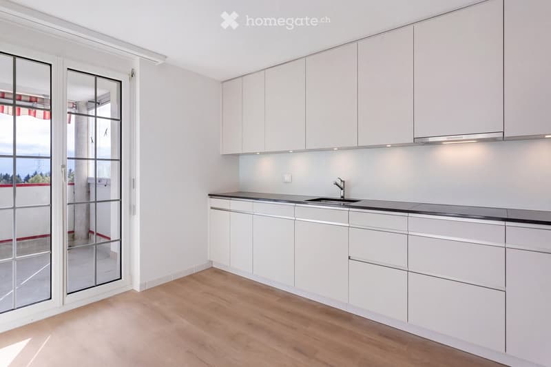 Modern kitchen with white cabinets, a black countertop, and a sink. The room features light wooden flooring and large glass doors leading to a balcony.