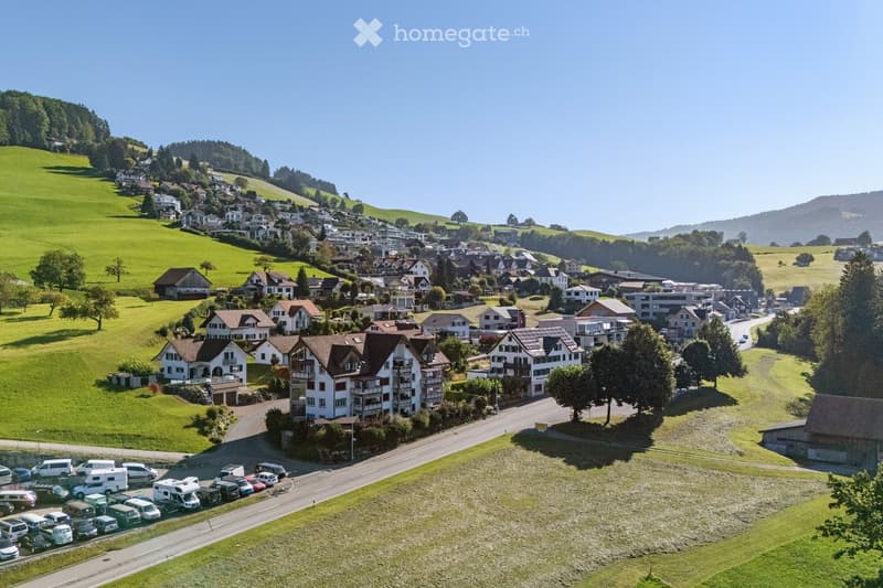 Aerial view of a small village with houses, green hills, and a parking area with several vehicles.