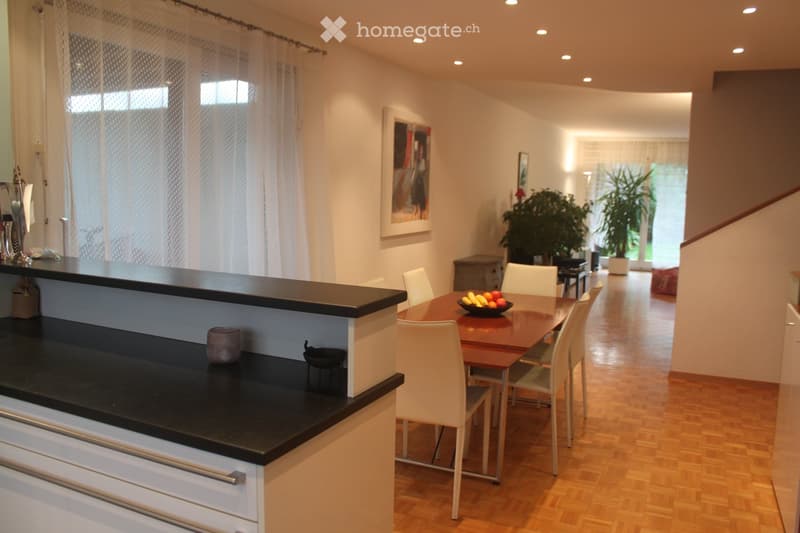 Modern kitchen and dining area with a black bar counter, white chairs, wooden dining table, and a bowl of fruit. The space features light wooden flooring, white walls, and large windows with sheer curtains.